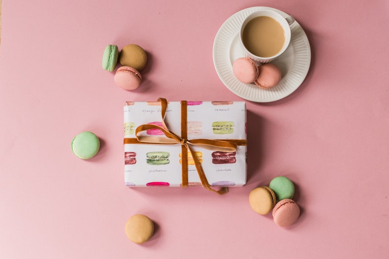 May include: A gift box wrapped in white paper with a brown ribbon and a pattern of colorful macarons. The box is surrounded by macarons in various colors, including pink, green, and brown. A cup of coffee and a saucer are in the upper right corner of the image.