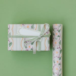 May include: A rectangular gift box wrapped in white paper with vertical light green stripes and a floral pattern of pink flowers and green leaves, tied with a light green velvet ribbon and a blank white rectangular gift tag, next to a paper roll.