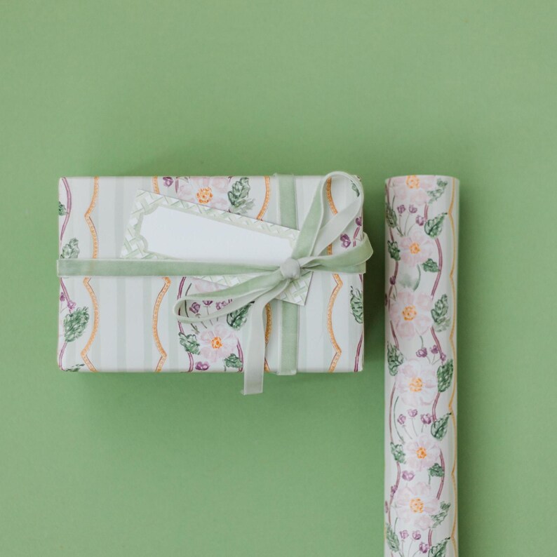 May include: A rectangular gift box wrapped in white paper with vertical light green stripes and a floral pattern of pink flowers and green leaves, tied with a light green velvet ribbon and a blank white rectangular gift tag, next to a paper roll.