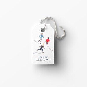 May include: A white gift tag with a watercolor illustration of four people ice skating. The text "Merry Christmas" is printed in blue at the bottom of the tag.