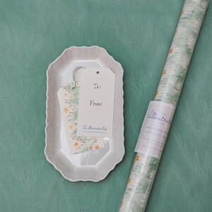 May include: A white ceramic tray holds gift tags and a small piece of patterned paper. A roll of wrapping paper with a green and white design and gold stars is next to the tray. The gift tags read "To:" and "From:" with the text "The Illustrated Life".