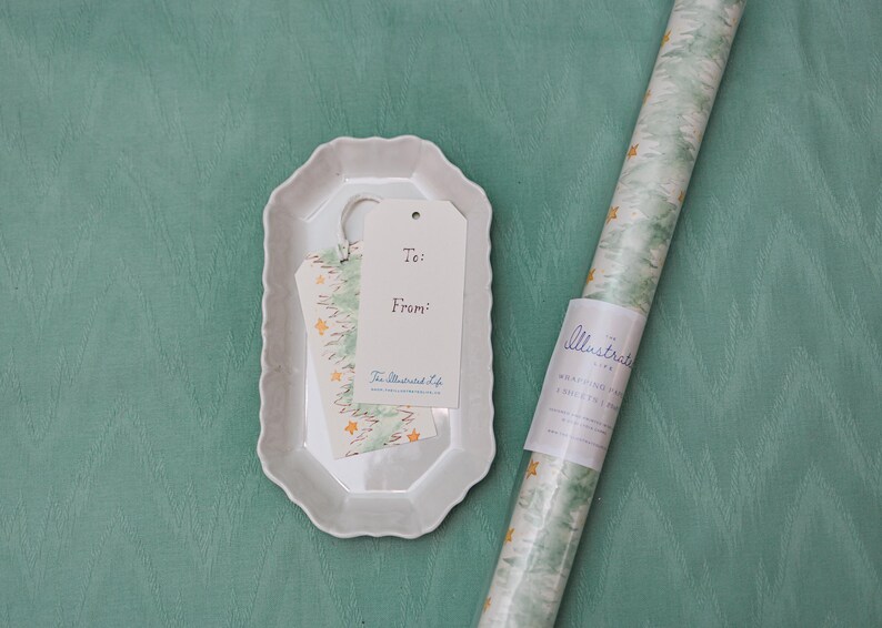 May include: A white ceramic tray holds gift tags and a small piece of patterned paper. A roll of wrapping paper with a green and white design and gold stars is next to the tray. The gift tags read "To:" and "From:" with the text "The Illustrated Life".