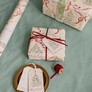 Puede incluir: Papel de regalo y etiquetas navide&ntilde;as. El papel de regalo presenta un estampado de &aacute;rboles de Navidad verdes, lazos rojos y estrellas doradas sobre un fondo blanquecino. Un regalo envuelto est&aacute; atado con cinta roja y una etiqueta que dice "Merry Christmas".