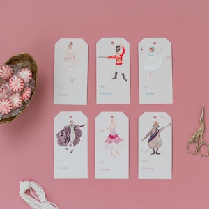 May include: Six illustrated gift tags featuring characters from the Nutcracker ballet. The tags are white with blue and pink accents. The characters include a ballerina, a soldier, a mouse king, and a sugar plum fairy. The tags are perfect for adding a festive touch to your gifts.