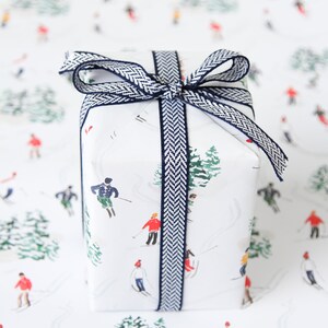 May include: A white gift box with a blue and white patterned ribbon. The box is wrapped in white paper with a repeating pattern of skiers and trees.
