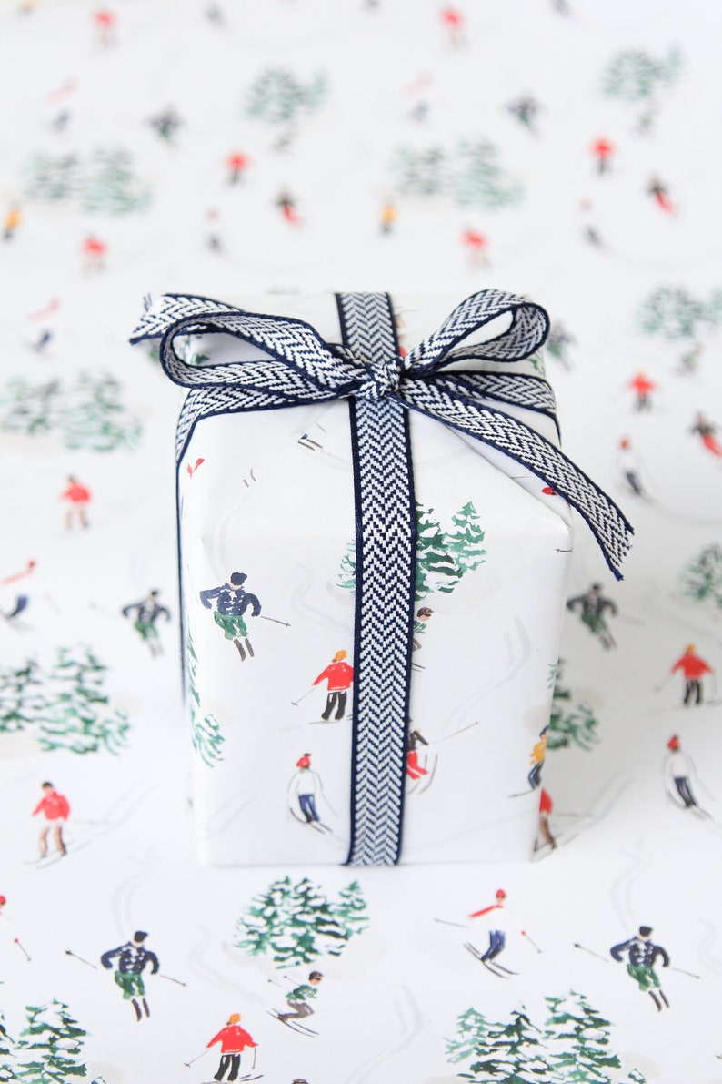 May include: A white gift box with a blue and white patterned ribbon. The box is wrapped in white paper with a repeating pattern of skiers and trees.