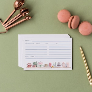May include: A stack of white recipe cards with blue text and lines, featuring a watercolor illustration of kitchen items at the bottom. The cards include fields for title, ingredients, serves, time, and directions, accompanied by copper measuring spoons, pink macarons, and a gold pen.