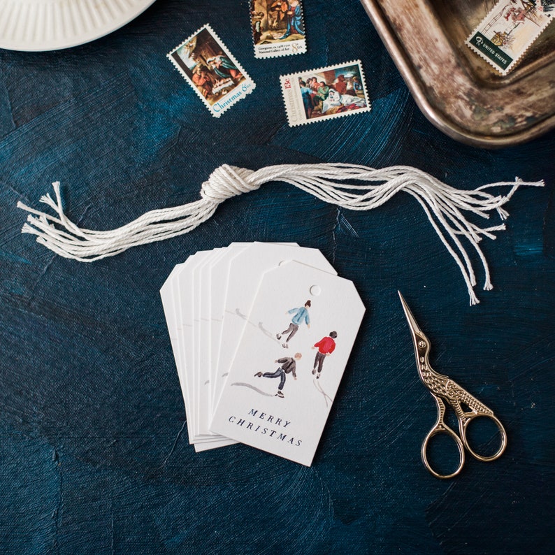 May include: A set of white gift tags with a watercolor illustration of children ice skating. The tags have a blue background and the text "Merry Christmas" in black.
