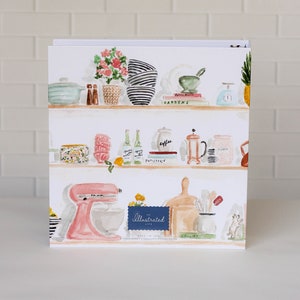 May include: A square, illustrated print featuring watercolor images of kitchen items on shelves. Items include a pink stand mixer, bowls, a mortar and pestle, and bottles. The text "Illustrated Life" is on a blue banner.