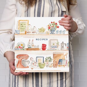May include: A square recipe book with a white cover featuring a watercolor illustration of three kitchen shelves filled with various items including a stand mixer, bowls, bottles, a vase of red flowers, and the word RECIPES printed in blue.