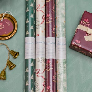 May include: An assortment of illustrated wrapping paper rolls in various festive designs, including Christmas trees, floral patterns, and ribbons. A wrapped gift box with a matching floral design is also present, along with decorative bells and a tag.