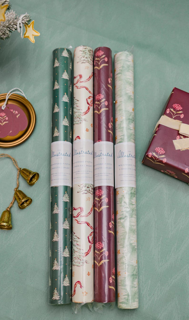 May include: An assortment of illustrated wrapping paper rolls in various festive designs, including Christmas trees, floral patterns, and ribbons. A wrapped gift box with a matching floral design is also present, along with decorative bells and a tag.