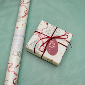 Puede incluir: Un rollo de papel de regalo ilustrado y una caja de regalo envuelta. Ambos est&aacute;n decorados con un patr&oacute;n repetido de &aacute;rboles de Navidad, cintas rojas y estrellas doradas sobre un fondo crema. La caja de regalo tiene una etiqueta burdeos.