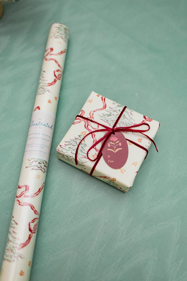 Puede incluir: Un rollo de papel de regalo ilustrado y una caja de regalo envuelta. Ambos est&aacute;n decorados con un patr&oacute;n repetido de &aacute;rboles de Navidad, cintas rojas y estrellas doradas sobre un fondo crema. La caja de regalo tiene una etiqueta burdeos.