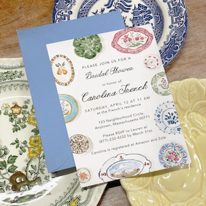 May include: A white bridal shower invitation card with a light blue envelope, featuring watercolor illustrations of various decorative plates. The text reads: Please join us for a Bridal Shower in honor of Carolina French, Saturday, April 12 at 11 AM.