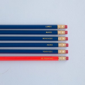 May include: A set of six pencils featuring five dark blue pencils labeled Lundi, Mardi, Mercredi, Jeudi, and Vendredi in gold lettering, and one red pencil labeled Le Weekend in gold lettering. Each pencil has a gold metal ferrule and a pink eraser.