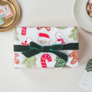 May include: A rectangular gift box wrapped in white paper featuring a repeating pattern of watercolor-style Christmas trees, candy canes, gingerbread people, snowmen, and Santa Claus faces, tied with a dark green velvet ribbon.