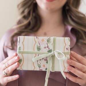 May include: A rectangular gift box wrapped in cream-colored paper featuring vertical light green stripes, gold wavy lines, and a floral pattern of pink flowers and green leaves, tied with a light sage green velvet ribbon in a bow.