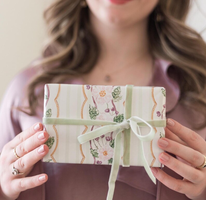May include: A rectangular gift box wrapped in cream-colored paper featuring vertical light green stripes, gold wavy lines, and a floral pattern of pink flowers and green leaves, tied with a light sage green velvet ribbon in a bow.