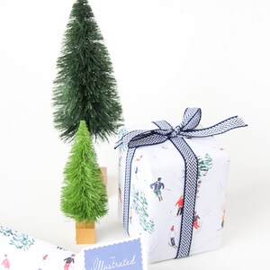 May include: White gift wrap with a blue and white ribbon. The wrap features a pattern of skiers on a snowy mountain. A small blue tag with the text "The Illustrated Life" is attached to the gift wrap. Two small green artificial Christmas trees are in the background.