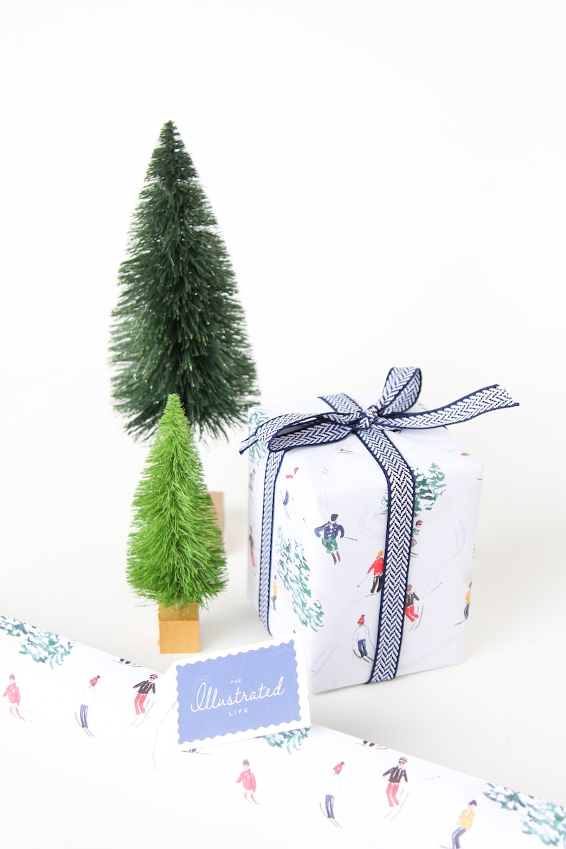 May include: White gift wrap with a blue and white ribbon. The wrap features a pattern of skiers on a snowy mountain. A small blue tag with the text "The Illustrated Life" is attached to the gift wrap. Two small green artificial Christmas trees are in the background.
