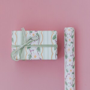 May include: A rectangular gift box wrapped in white paper with a pattern of pink flowers, green leaves, and gold wavy lines, tied with a light green velvet ribbon, alongside a matching roll of floral wrapping paper on a solid pink background.