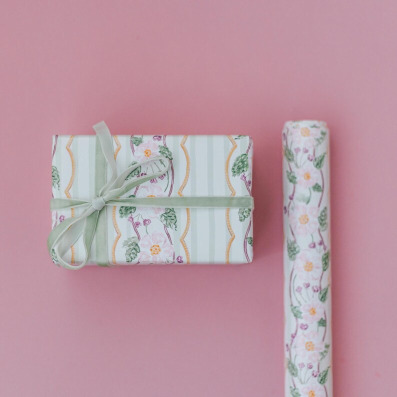 May include: A rectangular gift box wrapped in white paper with a pattern of pink flowers, green leaves, and gold wavy lines, tied with a light green velvet ribbon, alongside a matching roll of floral wrapping paper on a solid pink background.