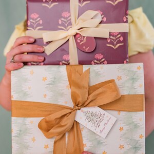May include: Two wrapped gifts are held in front of a person. The top gift is wrapped in burgundy paper with a floral pattern and tied with a cream ribbon. The bottom gift is wrapped in white paper with a tree and star pattern, tied with a gold ribbon, and a tag that says "Merry Christmas."
