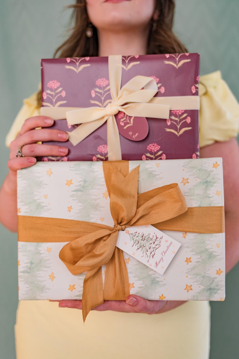 May include: Two wrapped gifts are held in front of a person. The top gift is wrapped in burgundy paper with a floral pattern and tied with a cream ribbon. The bottom gift is wrapped in white paper with a tree and star pattern, tied with a gold ribbon, and a tag that says "Merry Christmas."