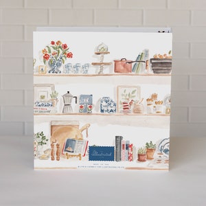 May include: A white three-ring binder featuring a watercolor illustration of kitchen shelves filled with various items including a vase of red flowers, a copper pot, a moka pot, books, and jars. The text "The Illustrated Life" is printed on the cover.