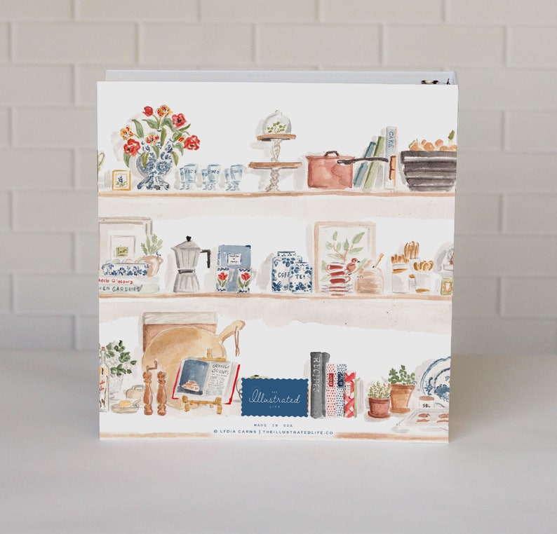 May include: A white three-ring binder featuring a watercolor illustration of kitchen shelves filled with various items including a vase of red flowers, a copper pot, a moka pot, books, and jars. The text "The Illustrated Life" is printed on the cover.