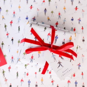 May include: A white gift box wrapped in paper with a pattern of people ice skating. The box is tied with a red ribbon and has a white gift tag that says "Merry Christmas".
