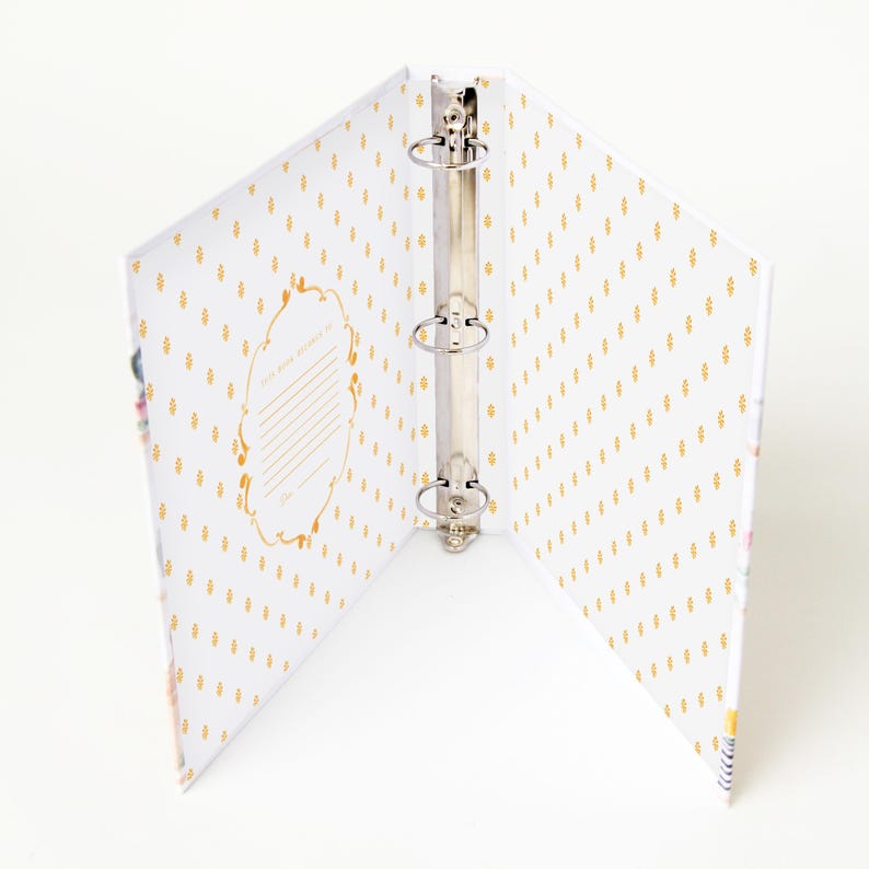 May include: Open white binder with a gold dotted pattern and a decorative gold frame. The binder has three silver rings and is designed to hold papers. The interior of the binder is white with a gold pattern.
