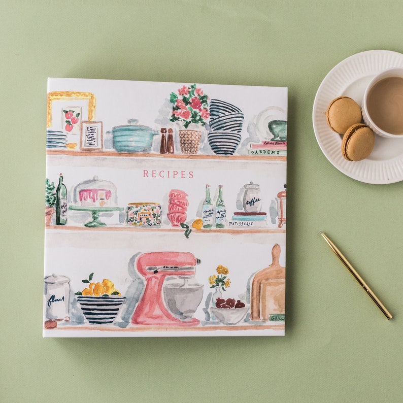 May include: A square recipe book with a watercolor illustration of kitchen shelves filled with cooking items. The word "RECIPES" is in pink letters. A plate with macarons and a cup of coffee are on the right.
