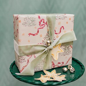 Puede incluir: Un regalo envuelto con papel de regalo festivo con &aacute;rboles de Navidad, lazos y estrellas. Est&aacute; atado con una cinta de terciopelo verde salvia y adornado con una etiqueta en forma de estrella y campanas doradas. El regalo est&aacute; sobre una bandeja de metal verde.
