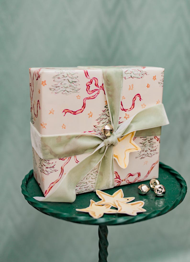 Puede incluir: Un regalo envuelto con papel de regalo festivo con &aacute;rboles de Navidad, lazos y estrellas. Est&aacute; atado con una cinta de terciopelo verde salvia y adornado con una etiqueta en forma de estrella y campanas doradas. El regalo est&aacute; sobre una bandeja de metal verde.