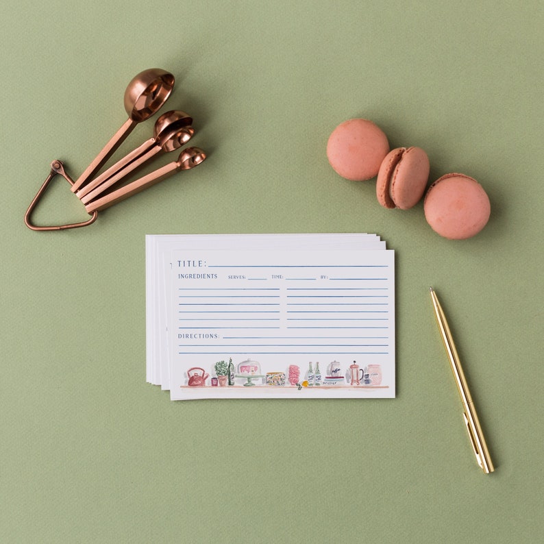 May include: A stack of white recipe cards with blue lines and text including Title, Ingredients, Serves, Time, By, and Directions, featuring a colorful kitchen illustration at the bottom, alongside copper measuring spoons, three pink macarons, and a gold pen.