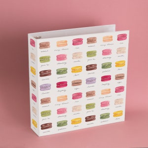 May include: A white binder with a colorful macaron pattern. Each macaron is labeled with a different flavor, including caramel, orange blossom, rose, green tea, strawberry, vanilla, chocolate, pistachio, lavender, raspberry, coffee, and more.