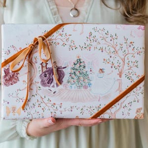 May include: A rectangular gift box wrapped in white paper featuring a watercolor illustration of a Christmas tree, a person in a purple cape, and a person in a white dress, tied with a mustard yellow velvet ribbon bow.