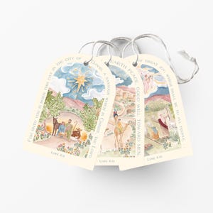 May include: Three arched paper gift tags with white string, featuring watercolor illustrations of the Nativity scene. The tags include text from Luke 2:10-14, depicting a star, a manger, camels, and shepherds in soft, muted colors on a cream background.