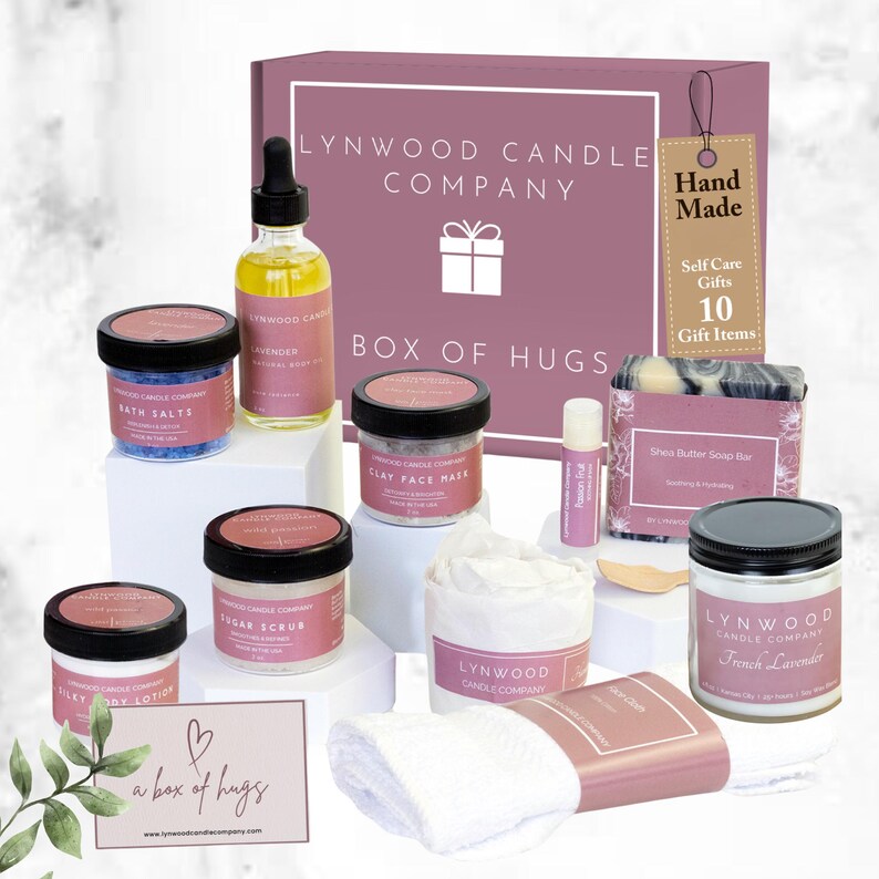 May include: A pink box with the text "Lynwood Candle Company" and "Box of Hugs" on the front. The box contains 10 self-care items, including a candle, bath salts, face mask, sugar scrub, body lotion, lip balm, soap bar, and a washcloth. The box is labeled "Hand Made" and "Self Care Gifts 10 Gift Items".