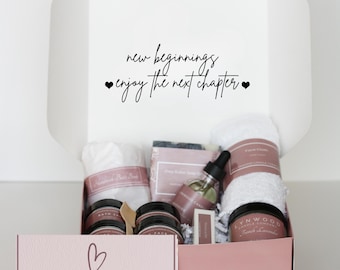 May include: A pink gift box with the text "new beginnings enjoy the next chapter" on the lid. The box is filled with bath and body products, including a white towel, a bar of soap, a bottle of oil, and three jars of cream. The bottom of the box has the text "a box of hugs" on it.