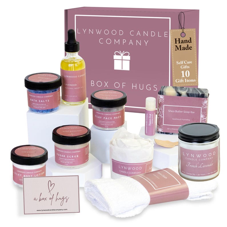 Pu&ograve; includere: Una scatola rosa con il testo "LYNWOOD CANDLE COMPANY" e "BOX OF HUGS" sul coperchio. La scatola contiene una variet&agrave; di articoli per la cura personale, tra cui sali da bagno, lozione per il corpo, scrub allo zucchero, maschera per il viso, una candela, una saponetta e un balsamo per le labbra. La scatola include anche un panno bianco e un biglietto che dice "a box of hugs".