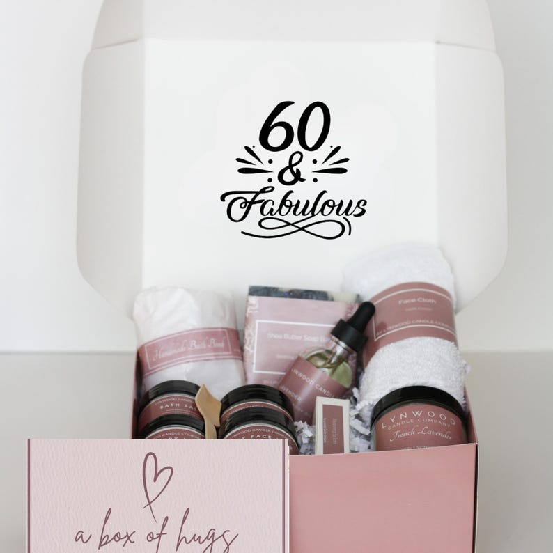 May include: A pink gift box with a white lid. The lid has a black graphic that says "60 & Fabulous". The box is filled with bath and body products, including a white towel, a bar of soap, a bottle of oil, and a jar of cream. The box also has a pink heart and the text "a box of hugs" on the front.