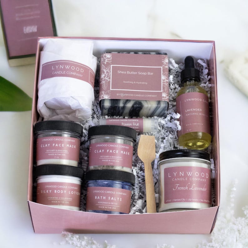 May include: A pink gift box filled with bath and body products from the Lynwood Candle Company. The box includes a candle, soap, lotion, bath salts, face mask, and essential oil. The products are all in white jars with black lids and feature a variety of scents, including lavender, French lavender, and passion fruit.