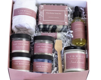 May include: A gift box filled with self-care products, including a candle, soap bar, bath salts, body lotion, face mask, and essential oil. The box is filled with shredded paper and the products are arranged neatly. The products are all from the same brand, Lynwood Candle Company.