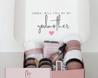 May include: A white gift box with the text "Emma, will you be my godmother" and a pink heart. Inside, a collection of self-care items, including bath products, a candle, and a bottle of oil. The box also contains a smaller pink box with the text "a box of hugs".