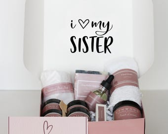 May include: A gift box with the text "i ♡ my SISTER" on the lid. Inside, a variety of self-care items are visible, including a white towel, jars, a bottle with a dropper, and a bar of soap. The box below reads "a box of hugs".