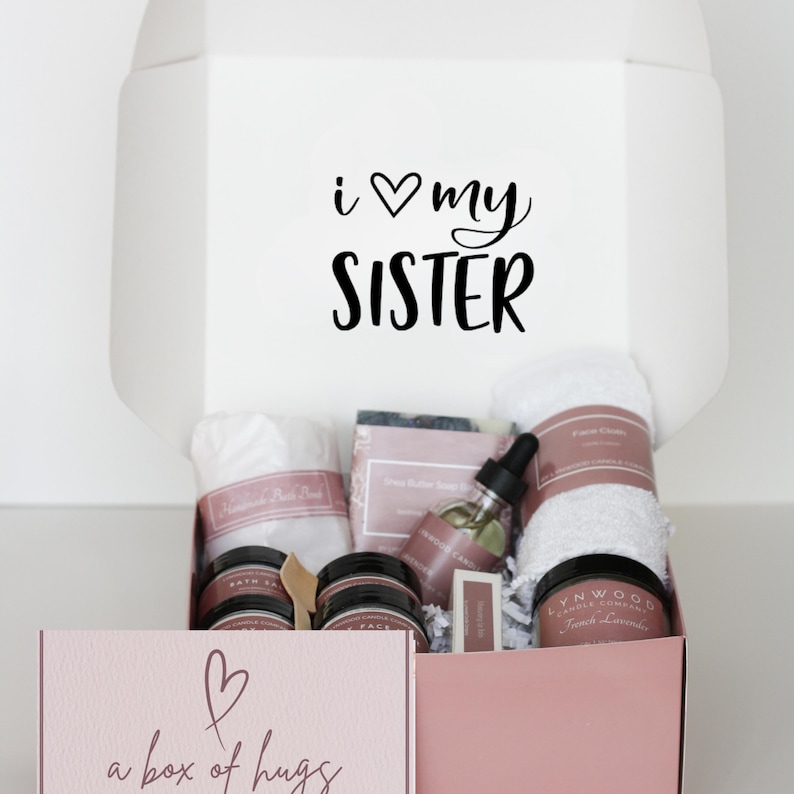 May include: A gift box with the text "i ♡ my SISTER" on the lid. Inside, a variety of self-care items are visible, including a white towel, jars, a bottle with a dropper, and a bar of soap. The box below reads "a box of hugs".
