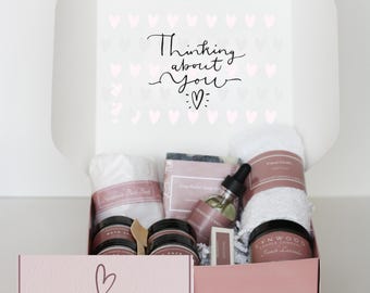 May include: A white gift box with a pink interior, filled with self-care items. The box includes a face cloth, candles, bath salts, soap, and a bottle of oil. The box lid has the text "Thinking about you" and the box front has the text "a box of hugs".